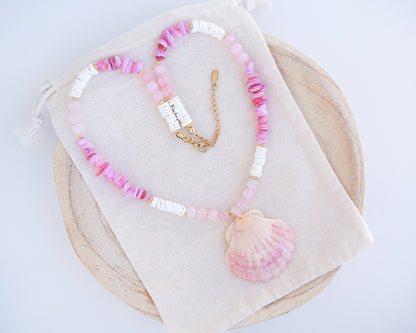 A necklace with pink and white beads and a shell pendant on light cotton displayed. A necklace with a big pink scallop shell paired with rose quartz gemstone beads, soft pink stones and white shell beads. The shell is a pilgrim's scallop shell handpicked from Portugal and is about 5 cm. The color of the shell is soft pink.