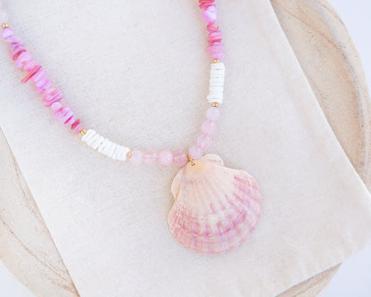 Close up of a necklace with pink and white beads and a shell pendant on light cotton displayed. A necklace with a big pink scallop shell paired with rose quartz gemstone beads, soft pink stones and white shell beads. The shell is a pilgrim's scallop shell handpicked from Portugal and is about 5 cm. The color of the shell is soft pink.