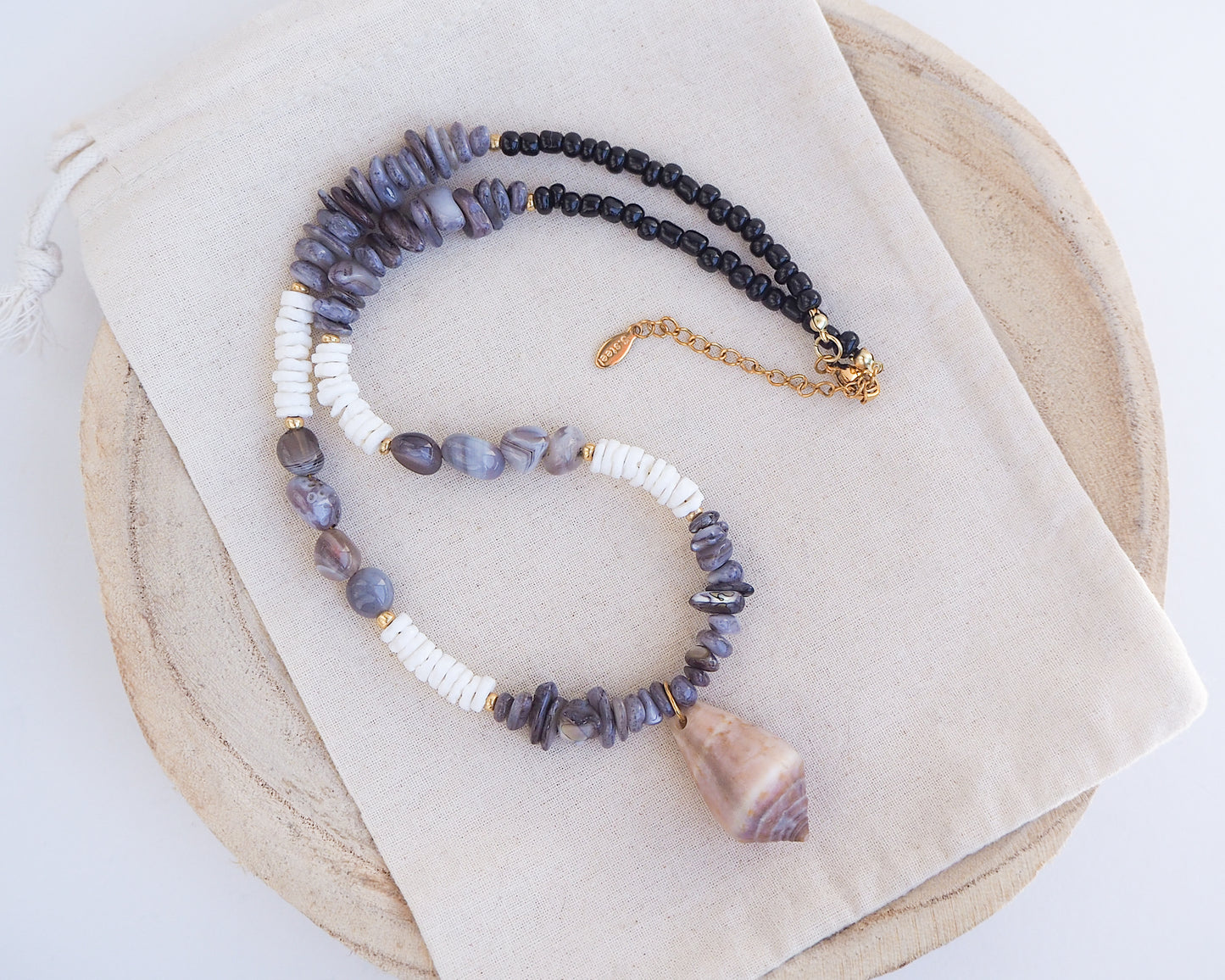 Front view of a necklace with grey and white beads and a large cone shell pendant on a light background. A necklace with a mediterranean cone shell paired with Botswana agate gemstone beads and white shell beads. The shell is a mediterranean cone shell from Portugal and is about 3,5 cm. The color of the shell is brown.
