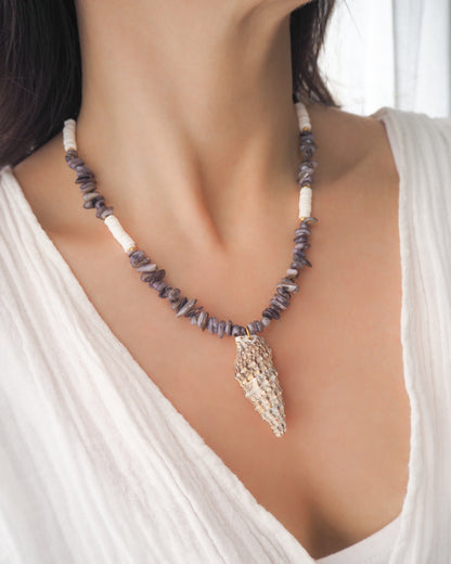 Model wearing a necklace with grey stone and white shell beads and a large horn shell pendant on a light background. A necklace with a mediterranean horn shell shell paired with grey stone beads and white shell beads. The shell is a mediterranean horn shell from Portugal and is about 5 cm. The color of the shell is white with dark pattern.