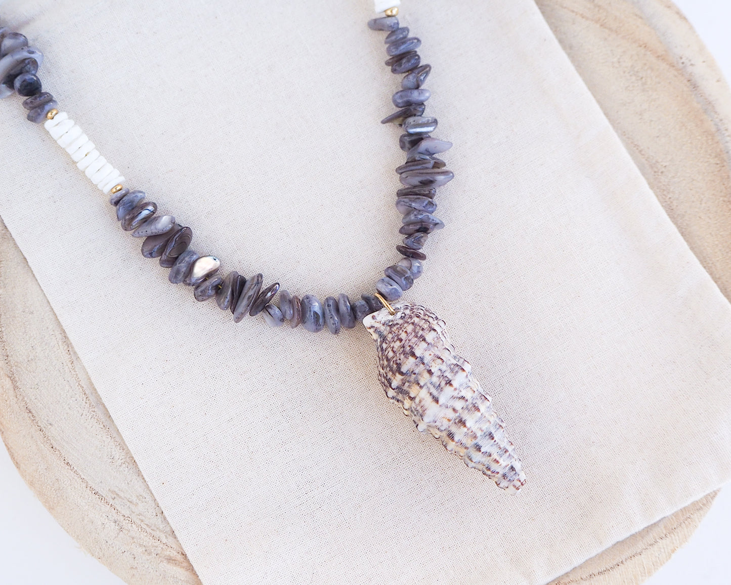 Close up of Necklace with grey stone and white shell beads and a large horn shell pendant on a light background. A necklace with a mediterranean horn shell shell paired with grey stone beads and white shell beads. The shell is a mediterranean horn shell from Portugal and is about 5 cm. The color of the shell is white with dark pattern.