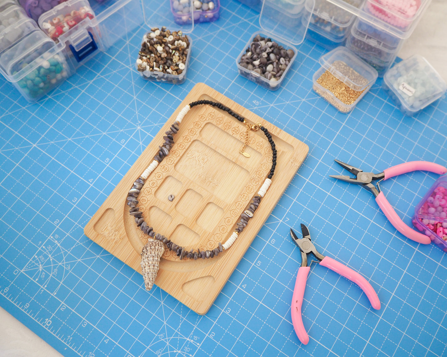 Jewelry-making setup with a necklace on a wooden board, pliers, and beads on a blue grid surface. Necklace with grey stone and white shell beads and a large horn shell pendant on a light background. A necklace with a mediterranean horn shell shell paired with grey stone beads and white shell beads. The shell is a mediterranean horn shell from Portugal and is about 5 cm. The color of the shell is white with dark pattern.
