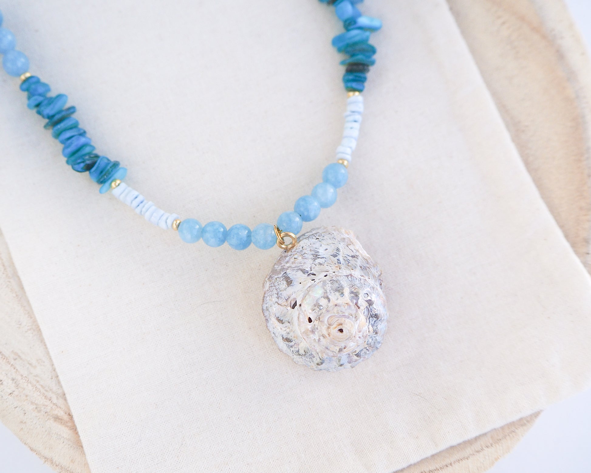 Close up of a necklace with a big star shell paired with aquamarine gemstone beads, turquoise stones and light blue shell beads on light background. The shell is a rough star shell from Portugal and is about 4 cm. The color of the shell is ivory grey.