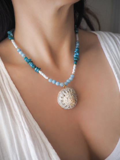 Model wearing a necklace with a big star shell paired with aquamarine gemstone beads, turquoise stones and light blue shell beads. The shell is a rough star shell from Portugal and is about 4 cm. The color of the shell is ivory grey.