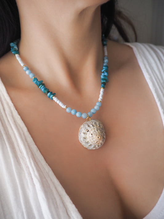 Model wearing a necklace with a big star shell paired with aquamarine gemstone beads, turquoise stones and light blue shell beads. The shell is a rough star shell from Portugal and is about 4 cm. The color of the shell is ivory grey.