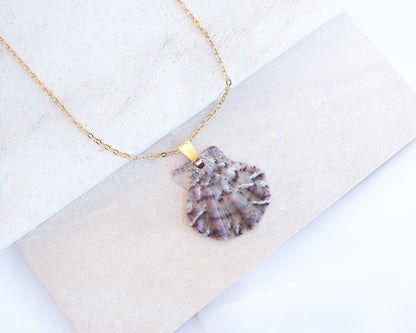 A gold chain necklace with a scallop shell pendant featuring natural grey purple, pink, and white marbled patterns, displayed on a light beige stone slab. The shell was found on the beaches of Portugal