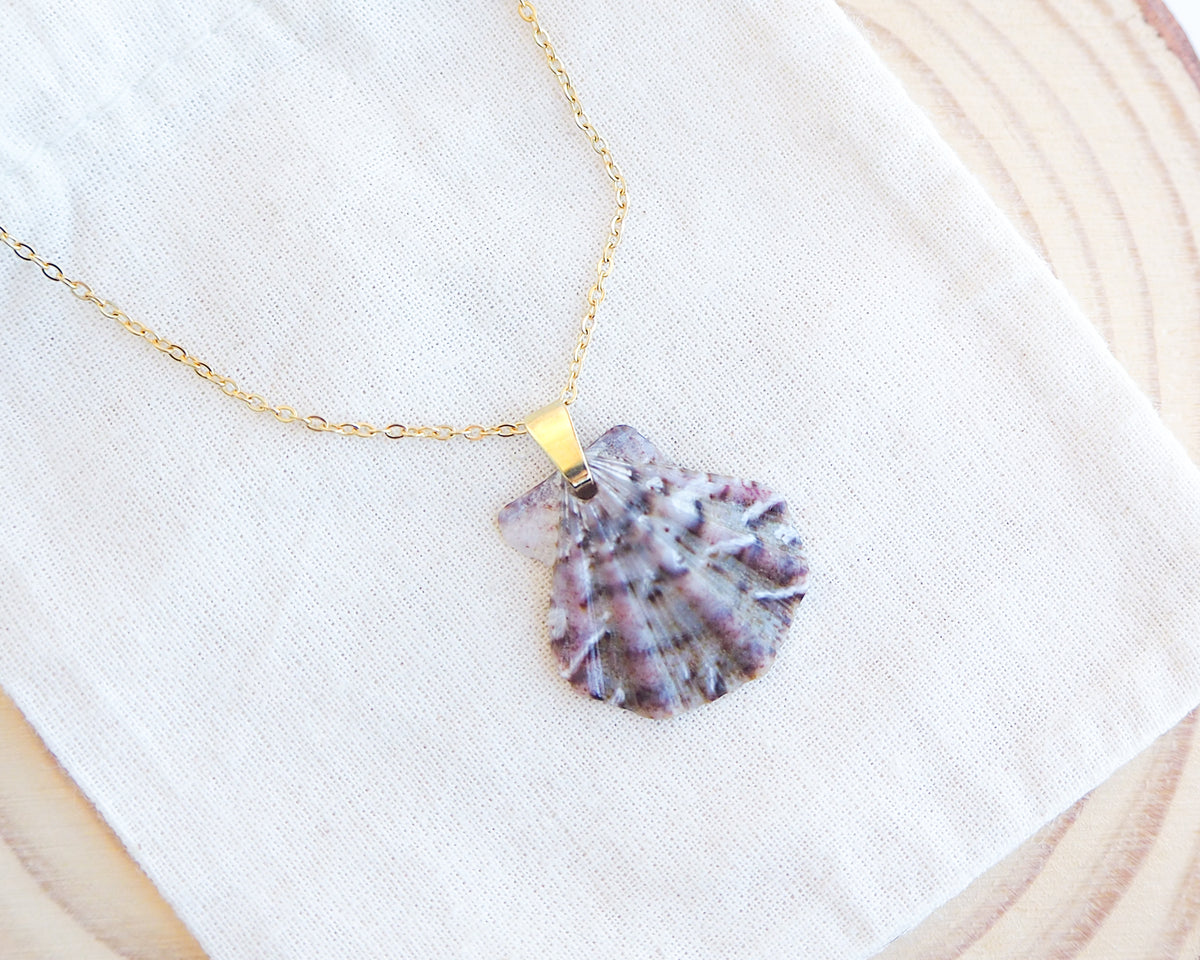 A gold chain necklace with a scallop shell pendant featuring purple and white marbled patterns, displayed on a textured white fabric over a wooden slice background.