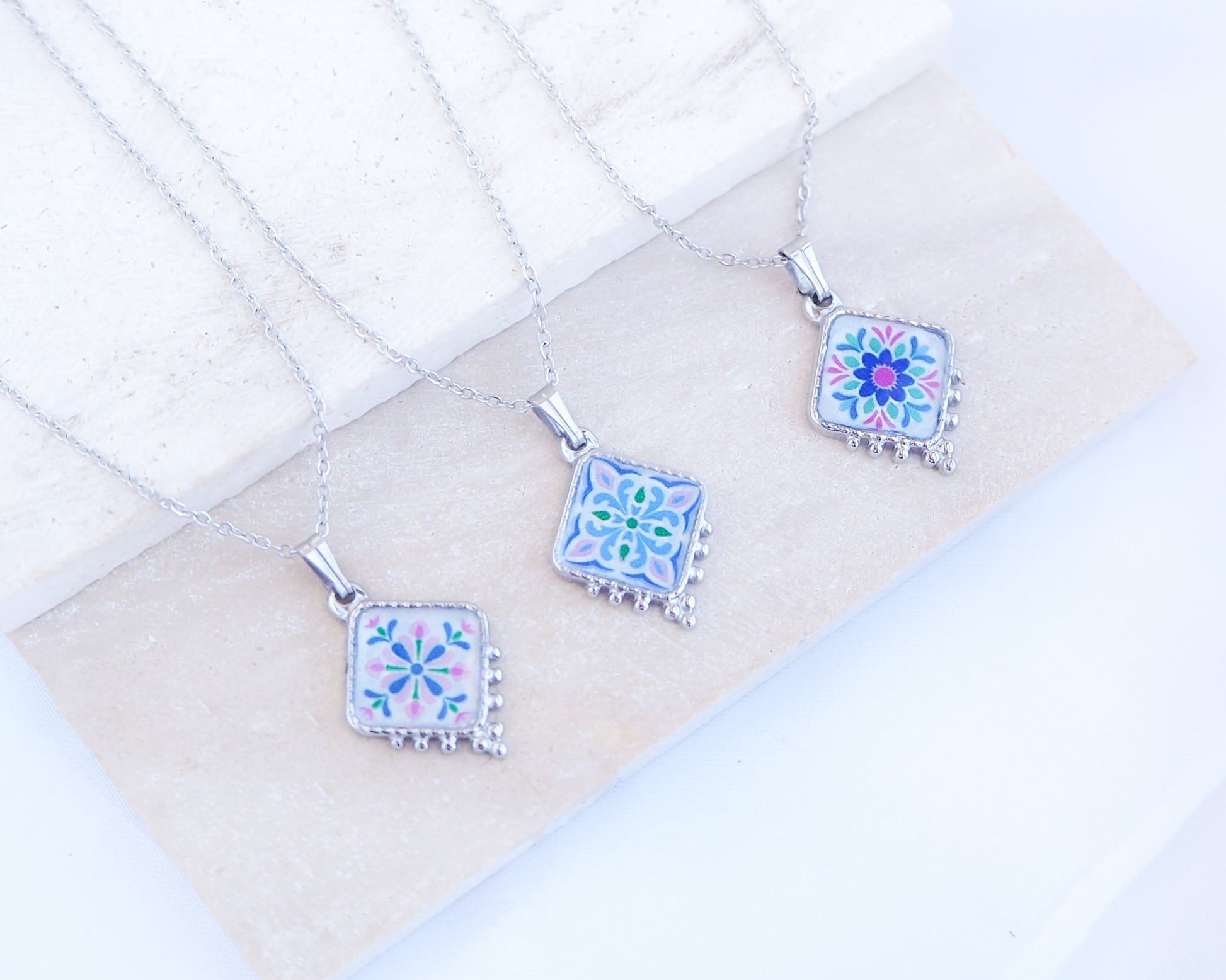Close up of 3 silver Portuguese Tile Necklace Floral Garden on stone displayed. Made with stainless steel or silver stainless steel and the colorful floral pendant are inspired by the Portuguese Azulejo design pattern. 