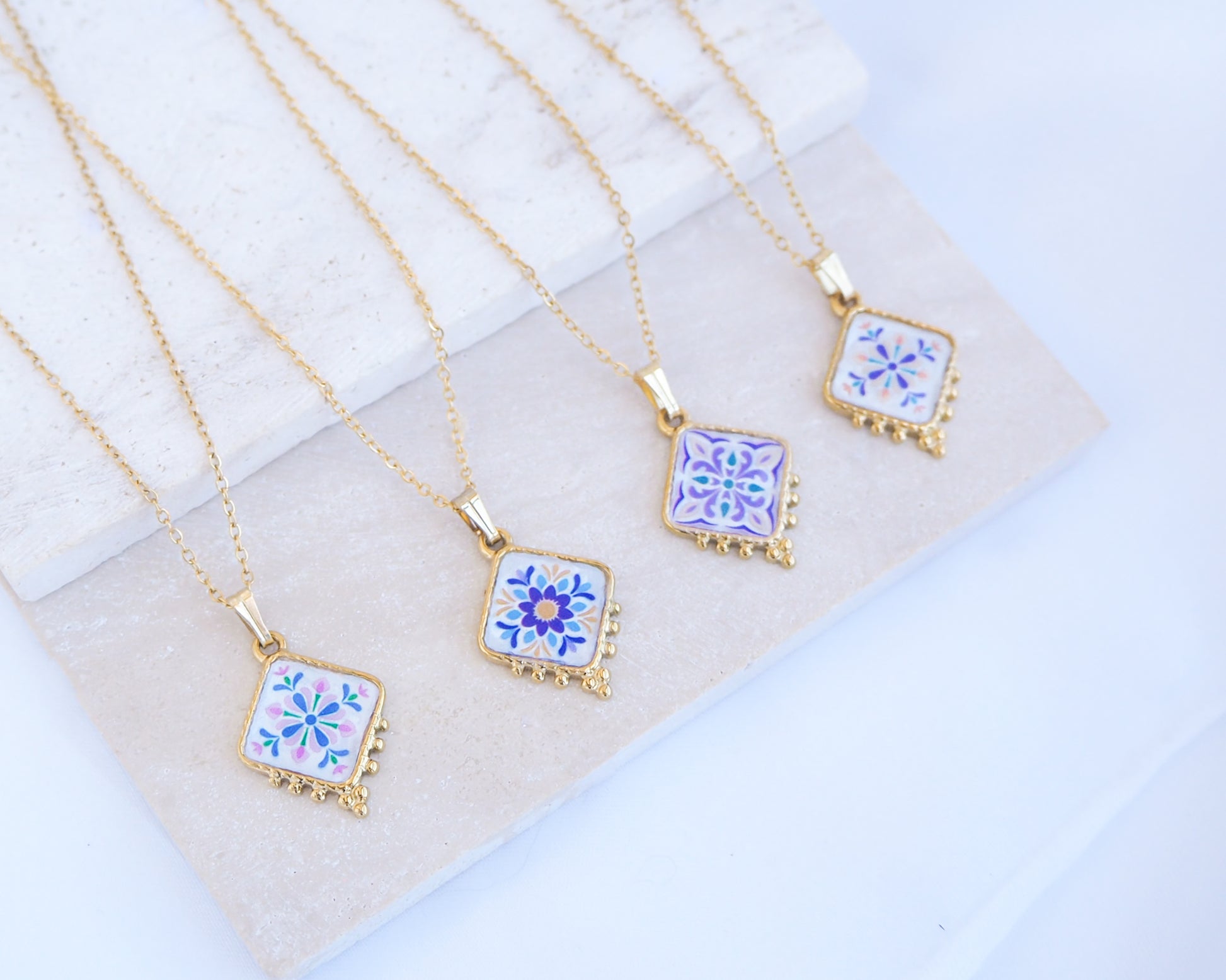 Four gold chain necklaces with square white enamel pendants decorated with blue and purple floral tile patterns, arranged on a light marble and beige stone block. Close up of 4 gold Portuguese Tile Necklace Floral Garden on stone displayed. Made with gold stainless steel and the colorful floral pendant are inspired by the Portuguese Azulejo design pattern. 