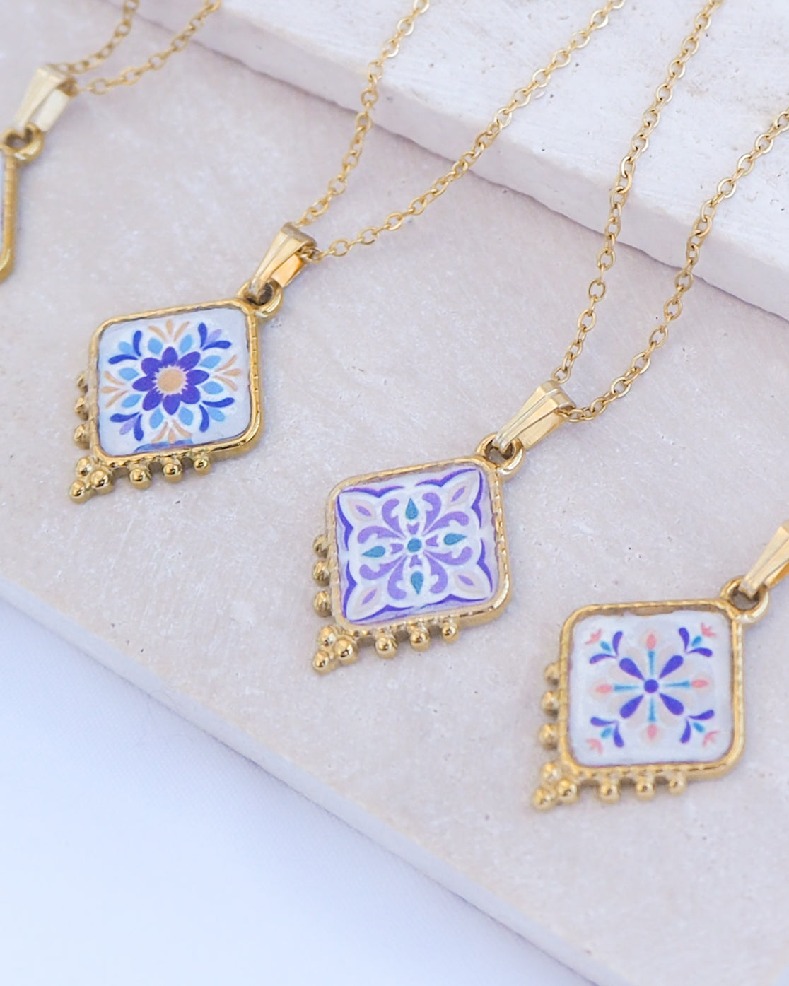 Three gold-tone necklaces with square enamel pendants featuring floral tile designs in blue, purple, and white, arranged on a light beige stone surface.