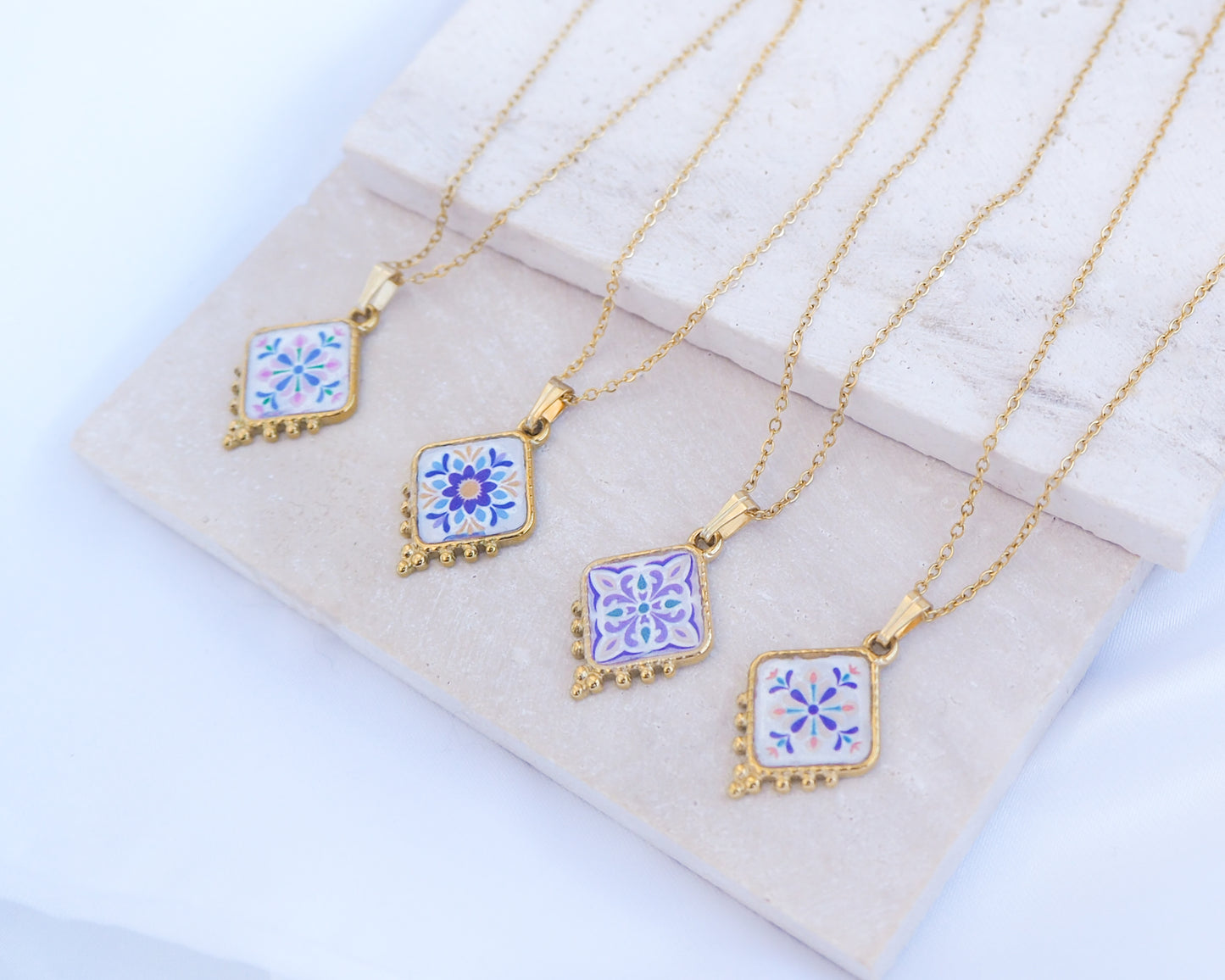Side view of 4 gold Portuguese Tile Necklace Floral Garden on stone displayed. Made with gold stainless steel and the colorful floral pendant are inspired by the Portuguese Azulejo design pattern. 