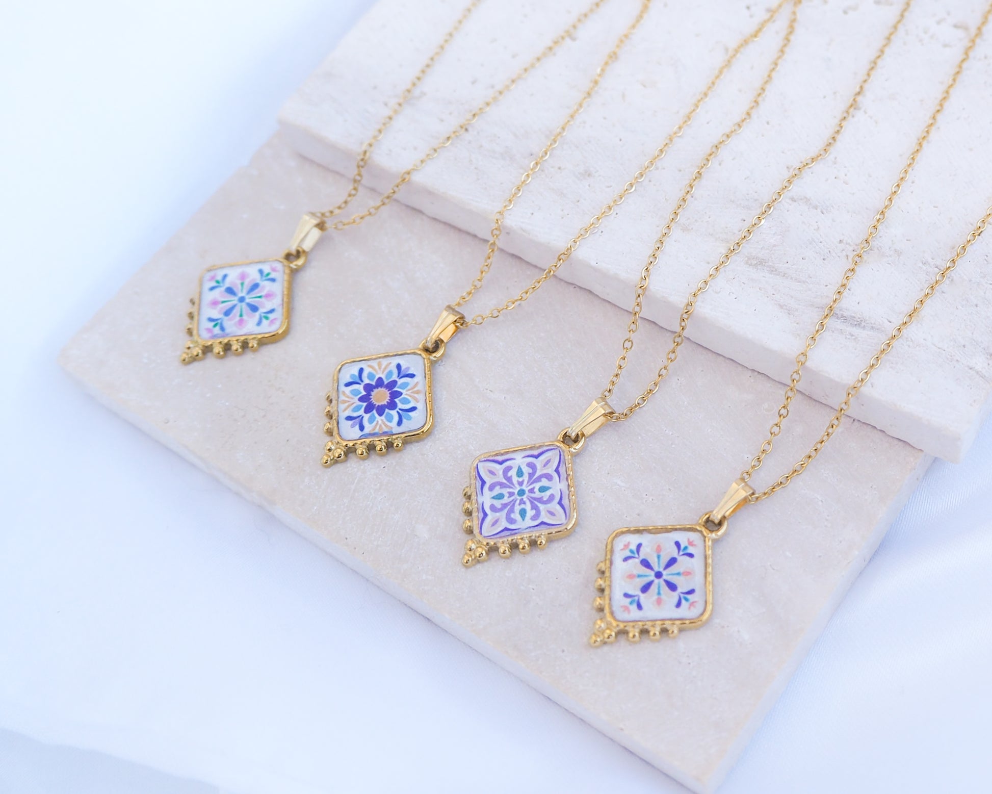 Side view of 4 gold Portuguese Tile Necklace Floral Garden on stone displayed. Made with gold stainless steel and the colorful floral pendant are inspired by the Portuguese Azulejo design pattern. 