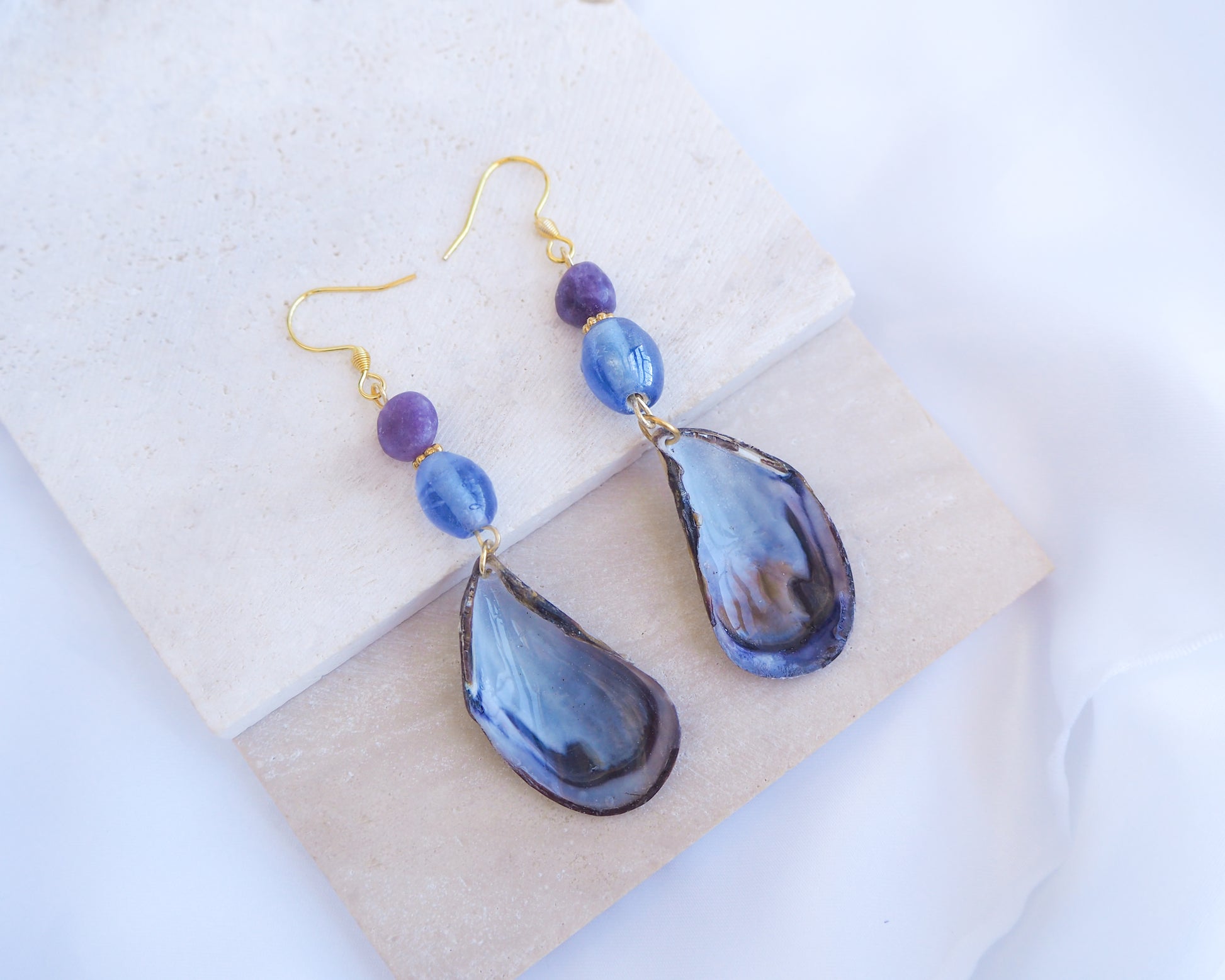 Stone displayed with handmade blue mussel shell earring with a gold earring hooks, purple amethyst gemstones and clear blue glass beads. The mussel shells were handpicked from the beaches of Portugal and they have a dark blue and white coloring.