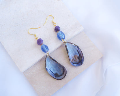 Stone displayed with handmade blue mussel shell earring with a gold earring hooks, purple amethyst gemstones and clear blue glass beads. The mussel shells were handpicked from the beaches of Portugal and they have a dark blue and white coloring.