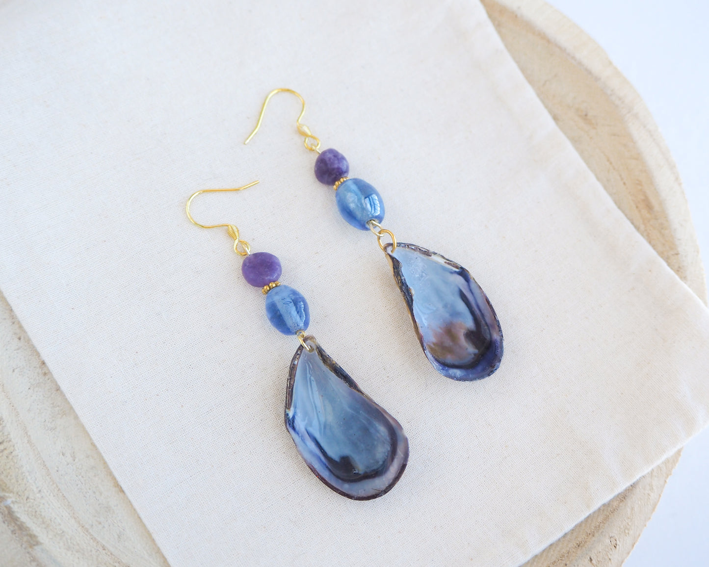 Handmade blue mussel shell earring with a gold earring hooks, purple amethyst gemstones and clear blue glass beads on cotton displayed. The mussel shells were handpicked from the beaches of Portugal and they have a dark blue and white coloring.