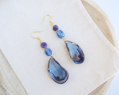 Handmade blue mussel shell earring with a gold earring hooks, purple amethyst gemstones and clear blue glass beads on cotton displayed. The mussel shells were handpicked from the beaches of Portugal and they have a dark blue and white coloring.