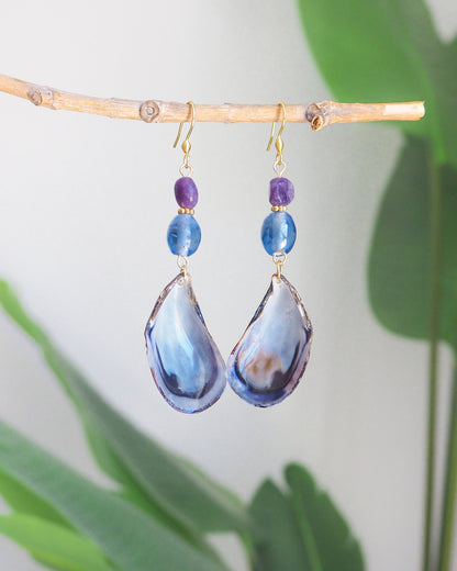 Front view of blue mussel shell earring with a gold earring hooks, purple amethyst gemstones and clear blue glass beads with green plant background. The mussel shells were handpicked from the beaches of Portugal and they have a dark blue and white coloring.