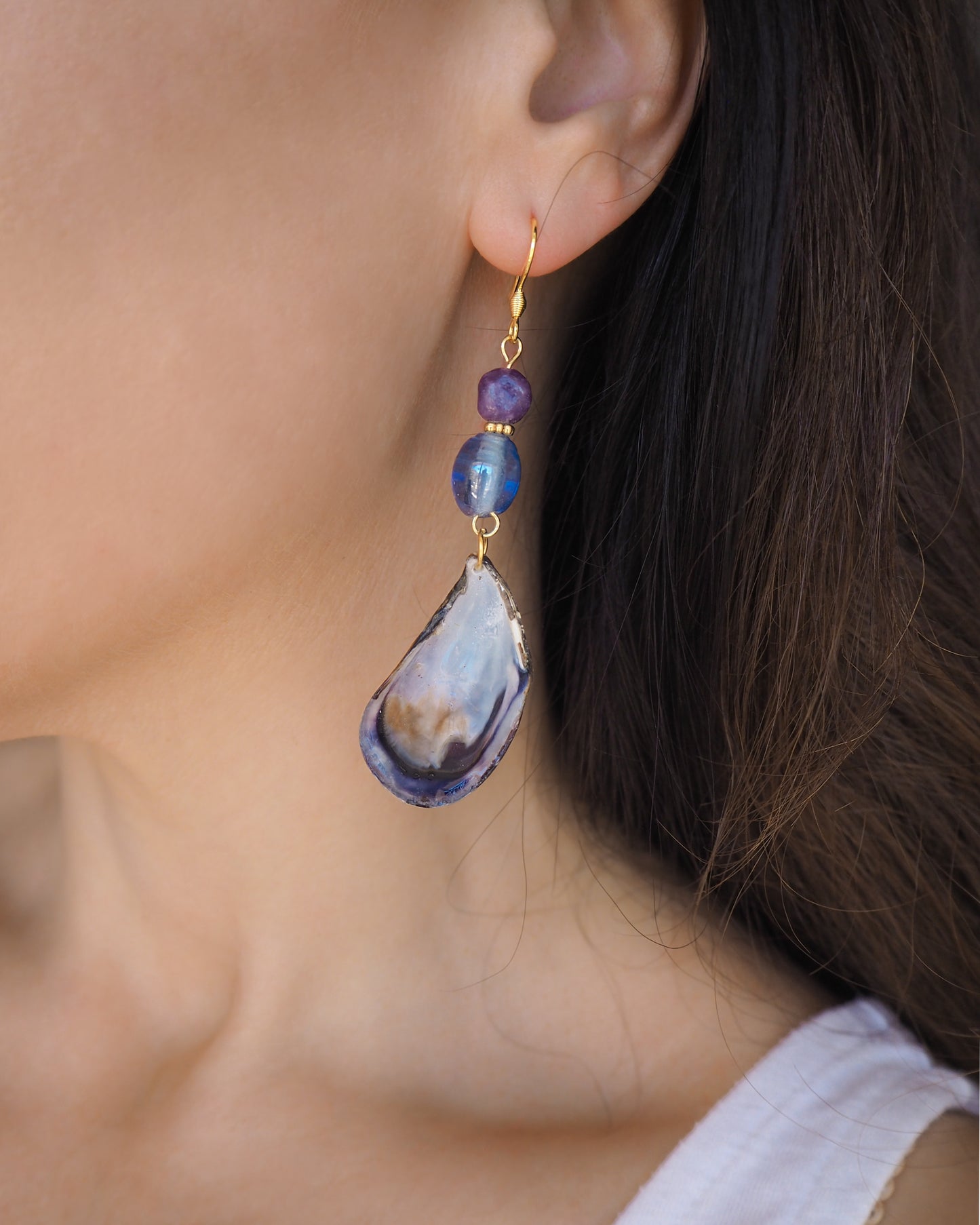 Model wearing handmade blue mussel shell earring with a gold earring hooks, purple amethyst gemstones and clear blue glass beads. The mussel shells were handpicked from the beaches of Portugal and they have a dark blue and white coloring.