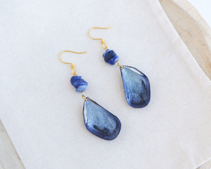 Blue mussel shell earring with a gold fishhook, a small dark blue lapis lazuli gemstones displayed on a light fabric. The mussel shells were handpicked from the beaches of Portugal and they have a dark blue and white coloring.