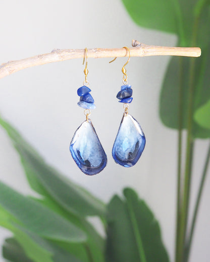 A pair of handmade dangle blue mussel shell earring with a gold fishhook, a small dark blue lapis lazuli gemstones. The mussel shells were handpicked from the beaches of Portugal and they have a dark blue and white coloring. The earrings are set in front of a green plant background.
