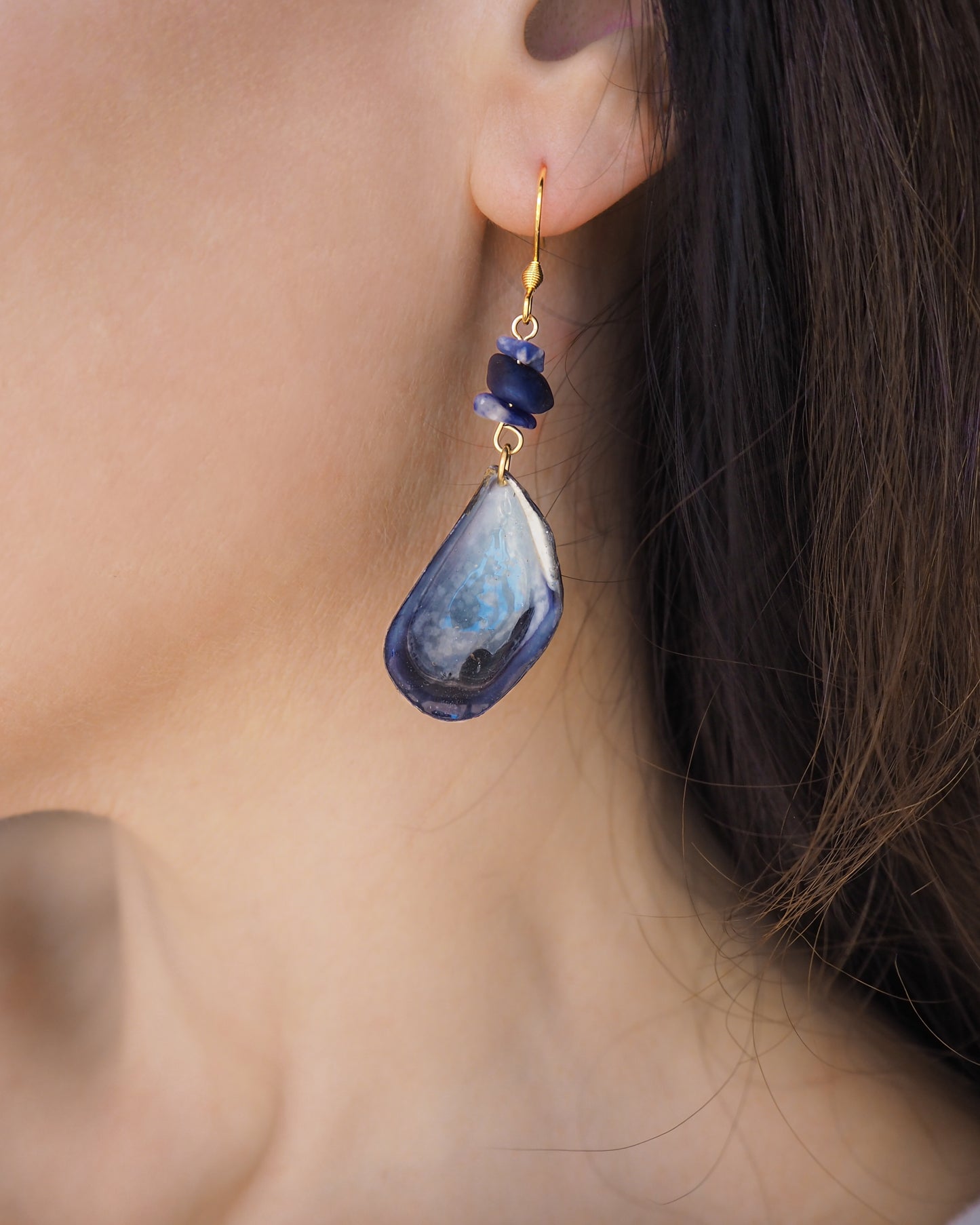 A close-up of a person's ear wearing a blue mussel shell earring with a gold fishhook, a small dark blue lapis lazuli gemstones. The mussel shells were handpicked from the beaches of Portugal and they have a dark blue and white coloring.
