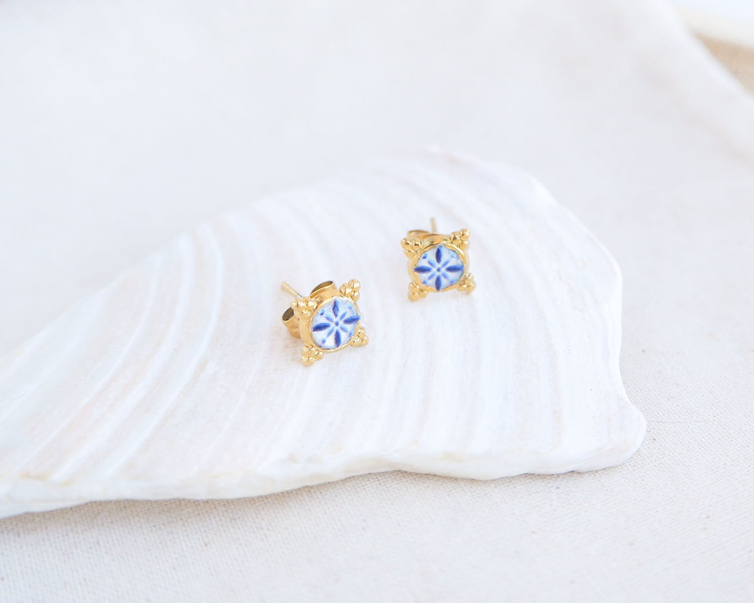 A pair of small gold stud earrings with blue and white portuguese tile pattern, displayed on a white seashell against a soft off-white background. Made with gold stainless steel and blue white center pattern that are inspired by the Portuguese Azulejo design pattern. 