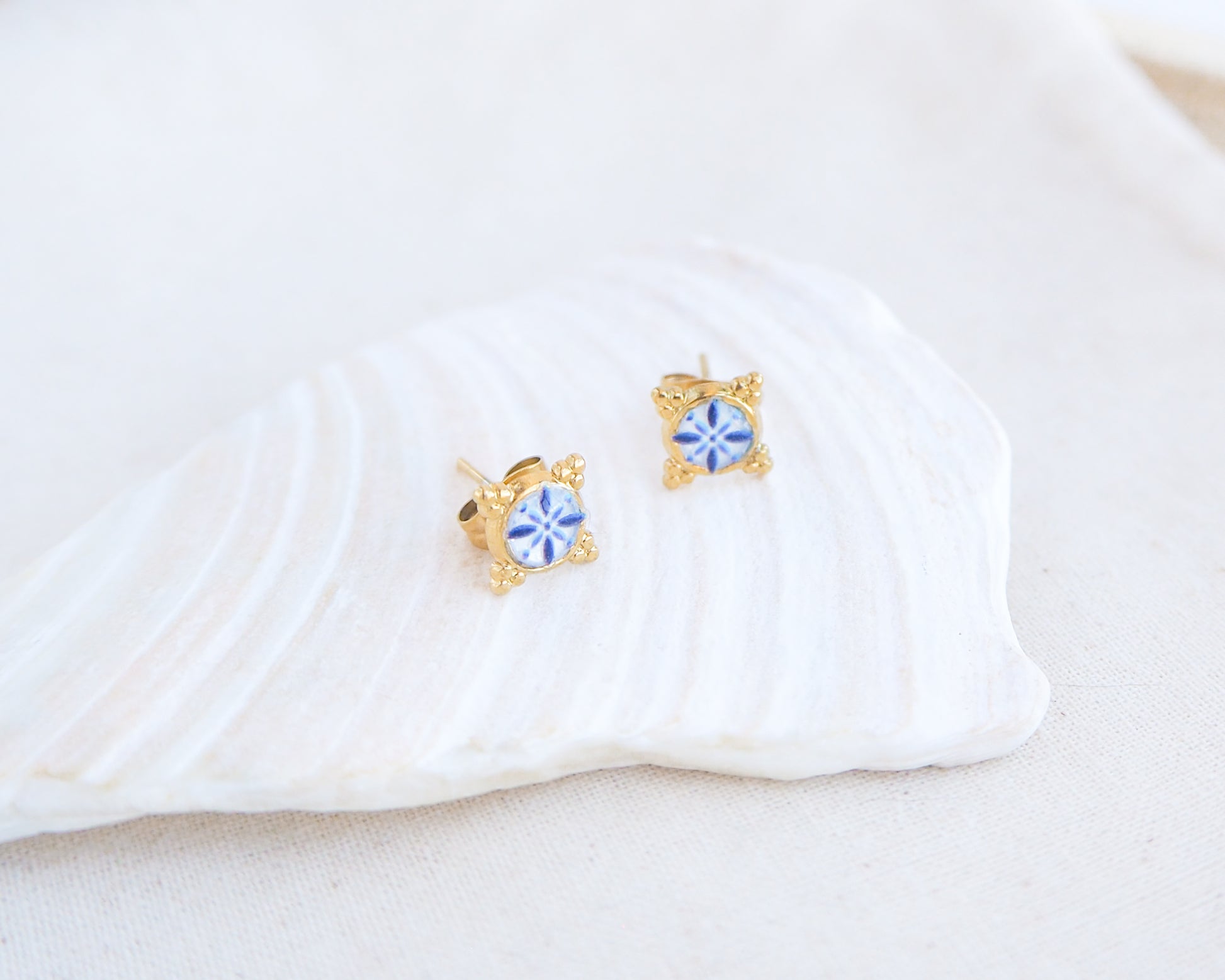 A pair of small gold stud earrings with blue and white portuguese tile pattern, displayed on a white seashell against a soft off-white background. Made with gold stainless steel and blue white center pattern that are inspired by the Portuguese Azulejo design pattern. 