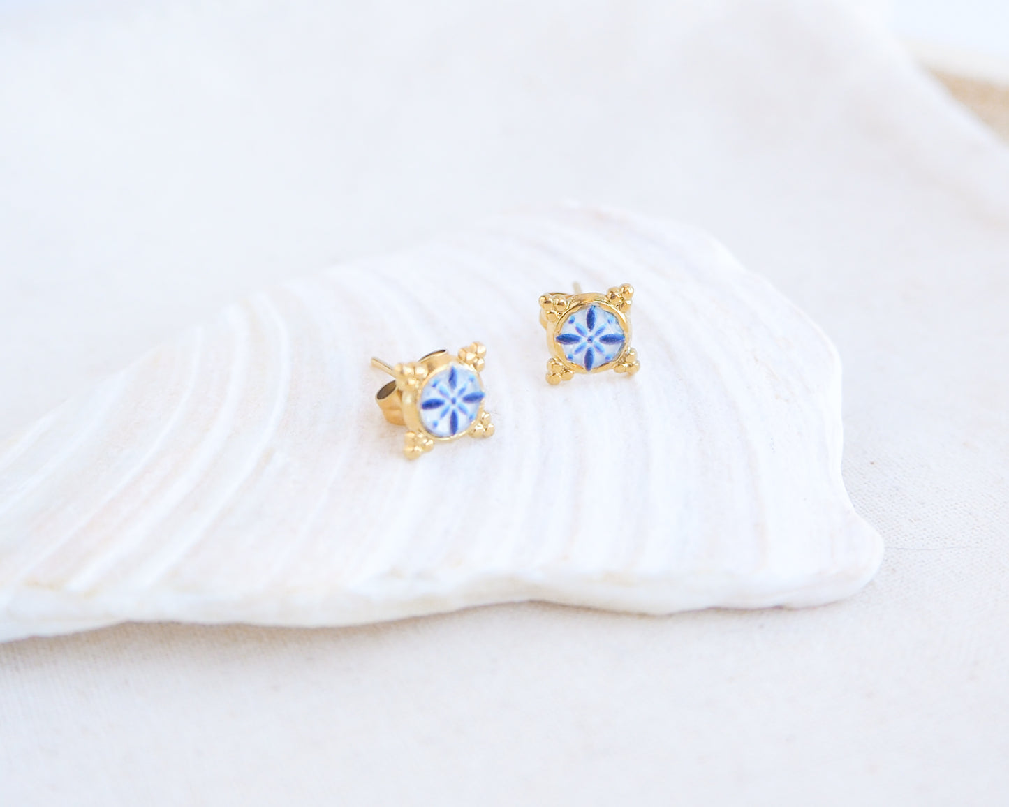 close up of a pair of small gold stud earrings with blue and white portuguese tile pattern, displayed on a white seashell against a soft off-white background. Made with gold stainless steel and blue white center pattern that are inspired by the Portuguese Azulejo design pattern. 