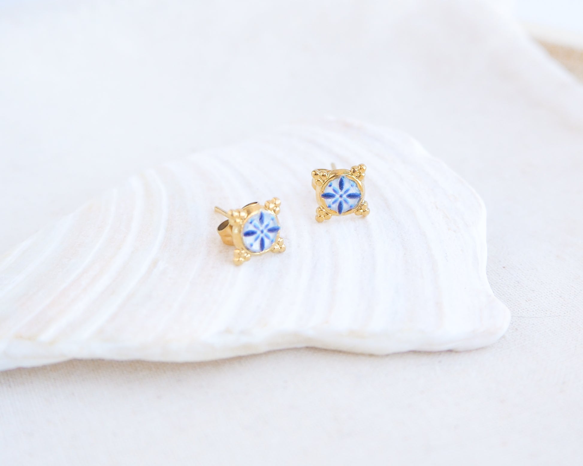 close up of a pair of small gold stud earrings with blue and white portuguese tile pattern, displayed on a white seashell against a soft off-white background. Made with gold stainless steel and blue white center pattern that are inspired by the Portuguese Azulejo design pattern. 