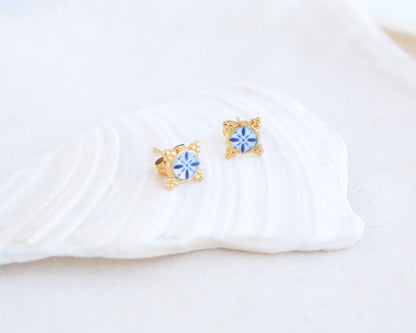 close up of a pair of small gold stud earrings with blue and white portuguese tile pattern, displayed on a white seashell against a soft off-white background. Made with gold stainless steel and blue white center pattern that are inspired by the Portuguese Azulejo design pattern. 