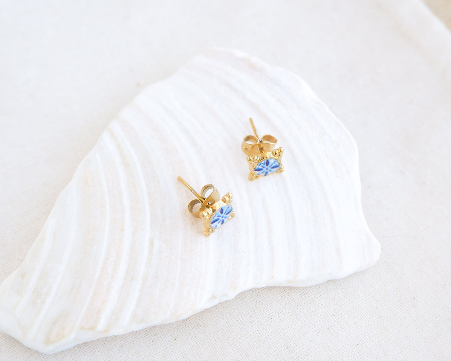 Front view of A pair of small gold stud earrings with blue and white Portuguese tile pattern, displayed on a white seashell against a soft off-white background. Made with gold stainless steel and blue white center pattern that are inspired by the Portuguese Azulejo design pattern. 
