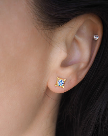 A close-up of a person's ear wearing a gold-toned stud earring with a blue and white starburst center, alongside a simple silver hoop earring. A pair of small gold stud earrings with blue and white Portuguese tile pattern, displayed on a white seashell against a soft off-white background. Made with gold stainless steel and blue white center pattern that are inspired by the Portuguese Azulejo design pattern. 