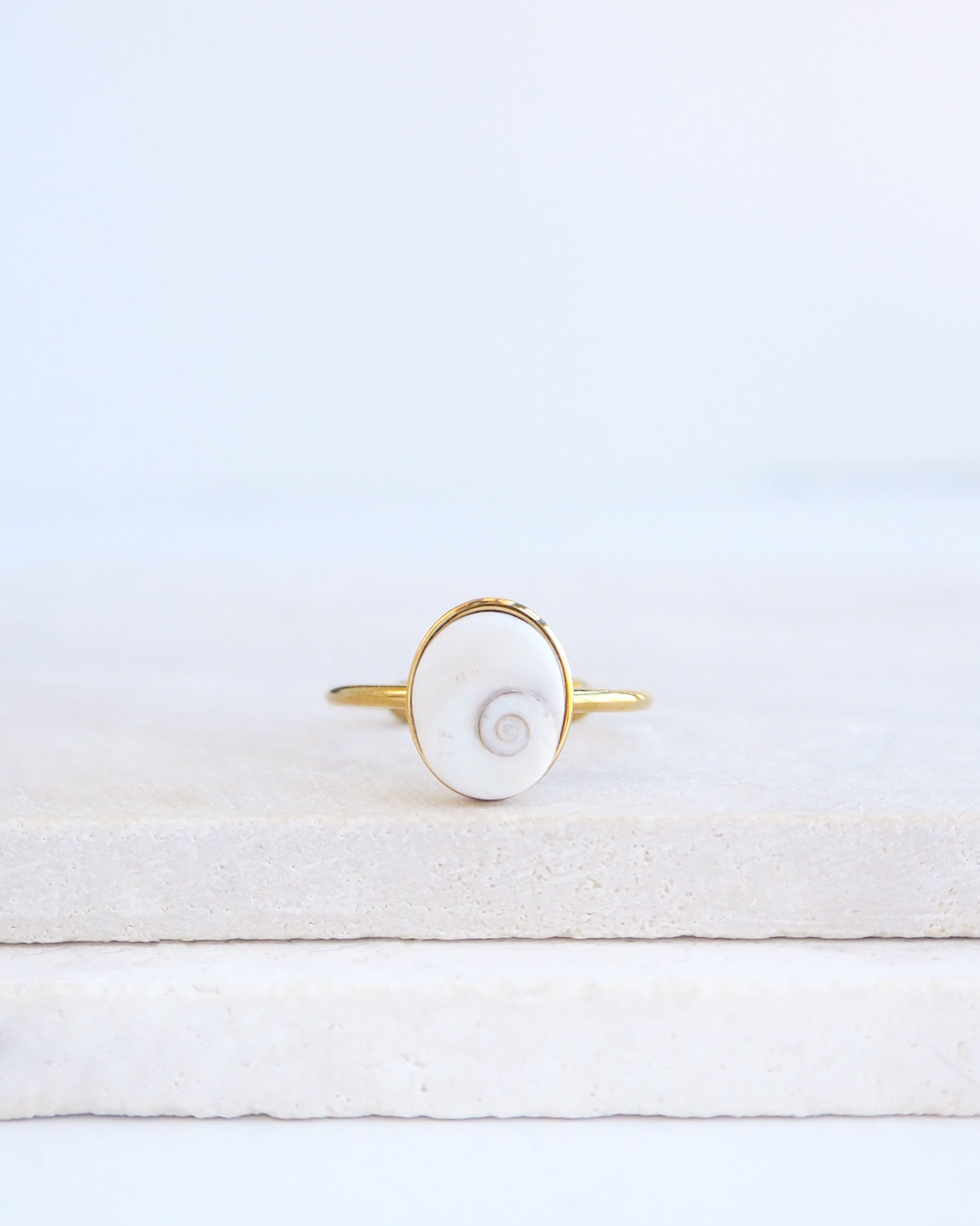 A gold ring featuring a white eye of saint lucia shiva eye shell inlay, displayed on a light textured stone surface against a soft white background.