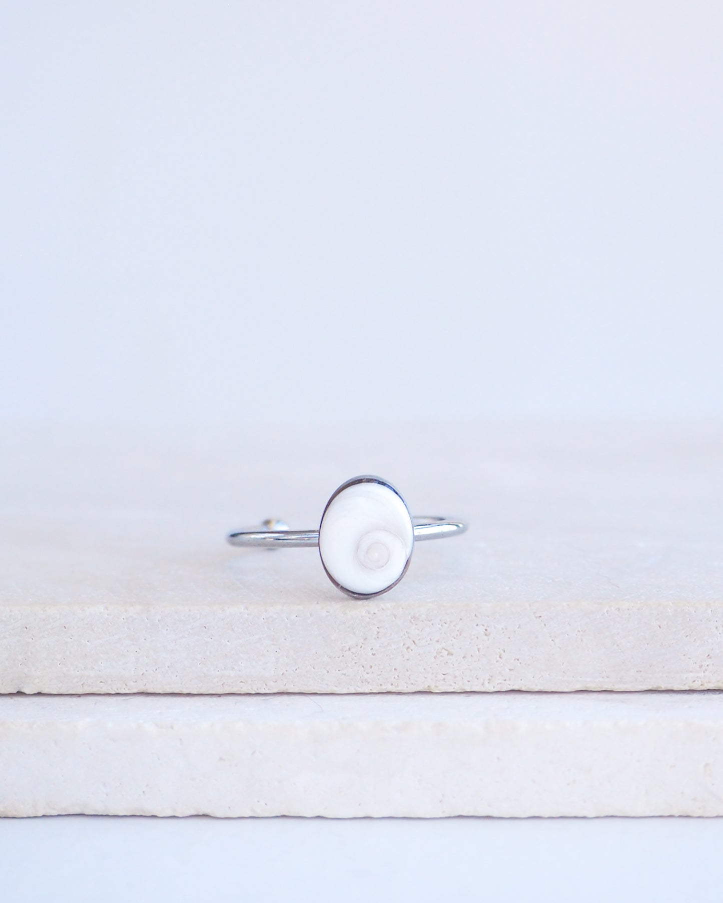 A silver ring with a round white eye of saint lucia shiva eye shell inlay, displayed on a light beige stone block against a white background.