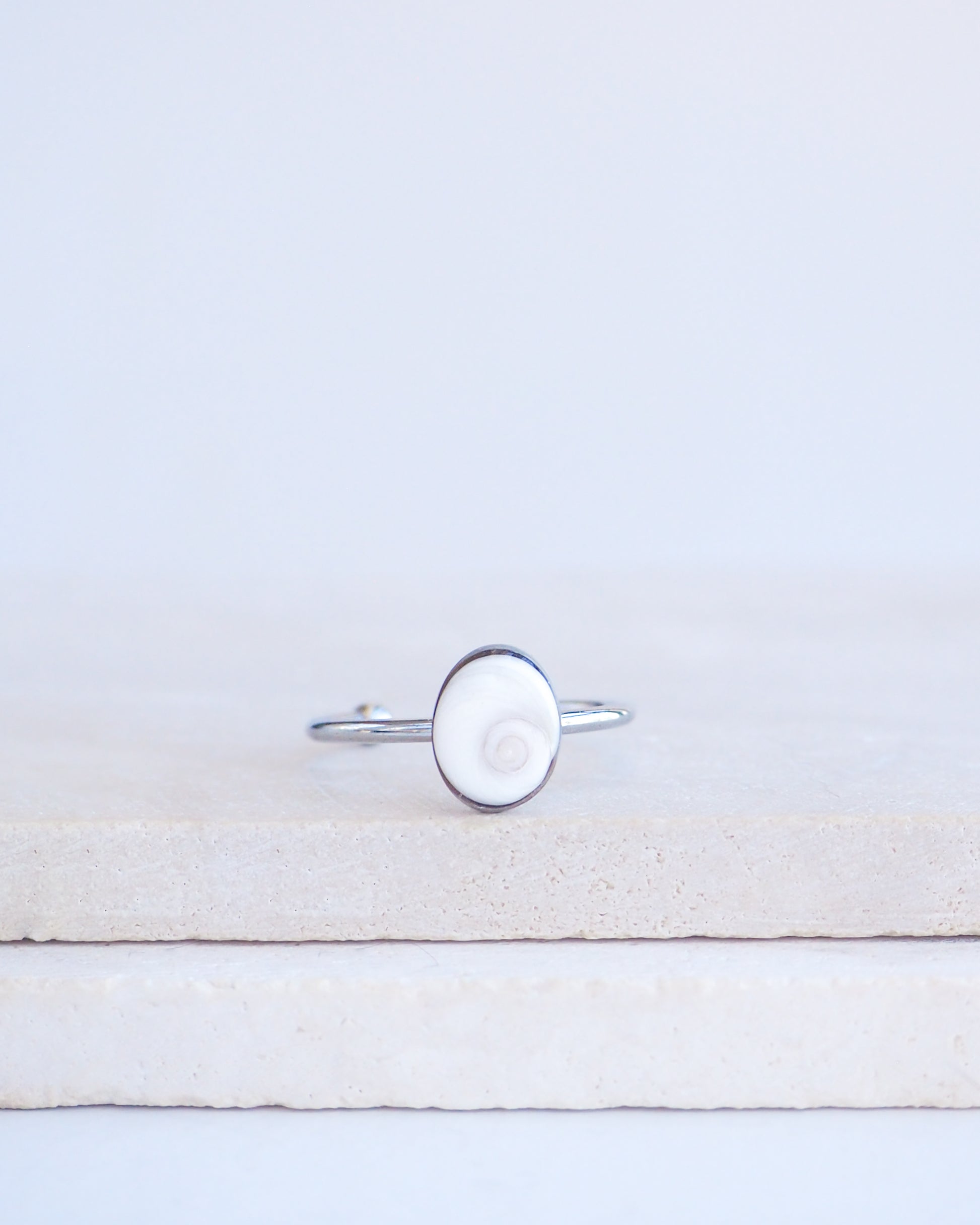 A silver ring with a round white eye of saint lucia shiva eye shell inlay, displayed on a light beige stone block against a white background.