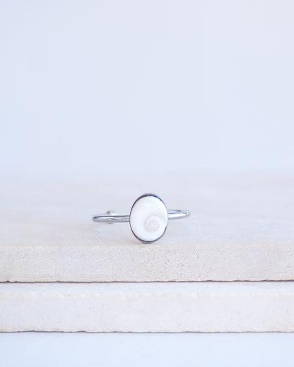 A silver ring with a round white eye of saint lucia shiva eye shell inlay, displayed on a light beige stone block against a white background.