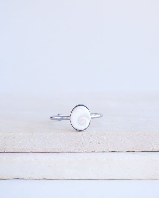 A silver ring with a round white eye of saint lucia shiva eye shell inlay, displayed on a light beige stone block against a white background.