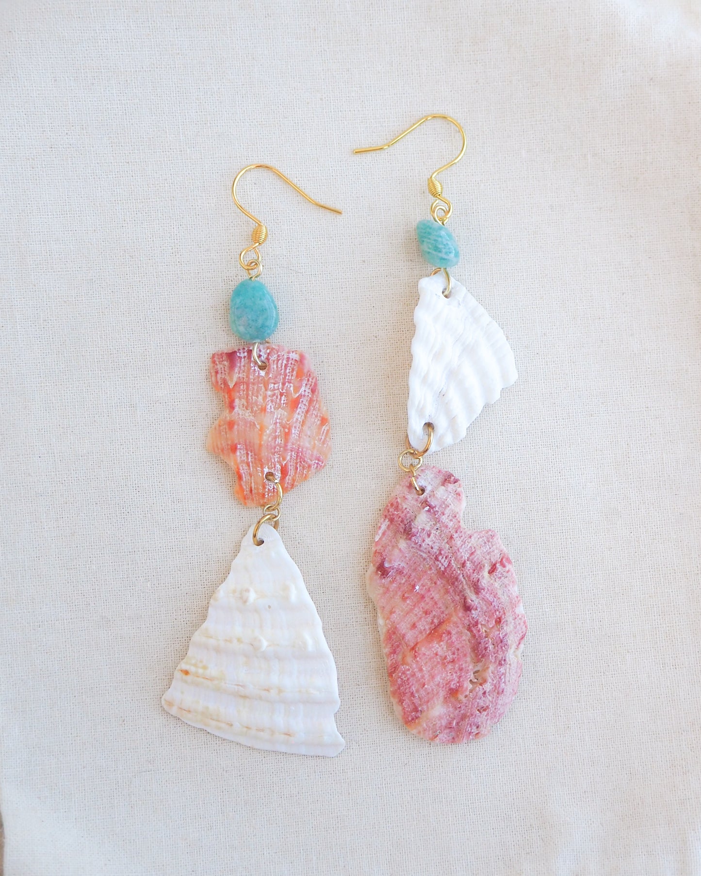 A pair of handmade dangle earrings featuring natural seashells in coral pink and white, accented with small turquoise beads and gold-tone hooks, displayed on a textured beige fabric background.