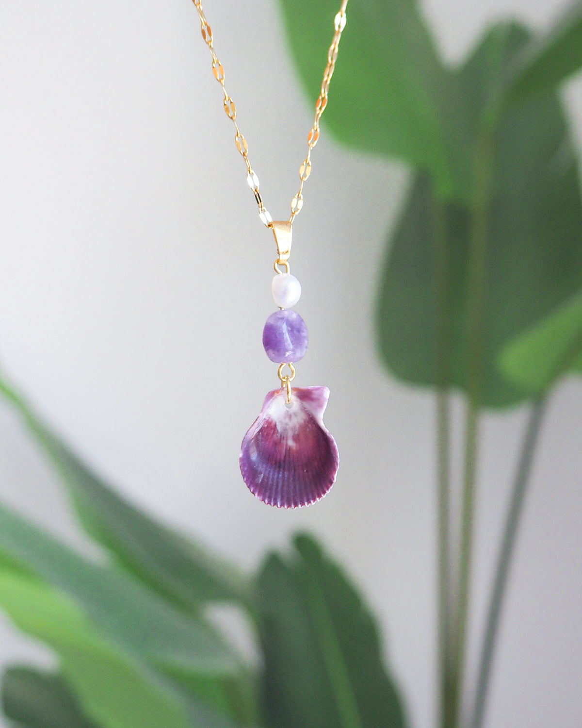 A gold chain necklace with a pendant featuring a purple seashell, a purple stone bead, and a small white pearl, shown against a soft-focus background of green leaves.