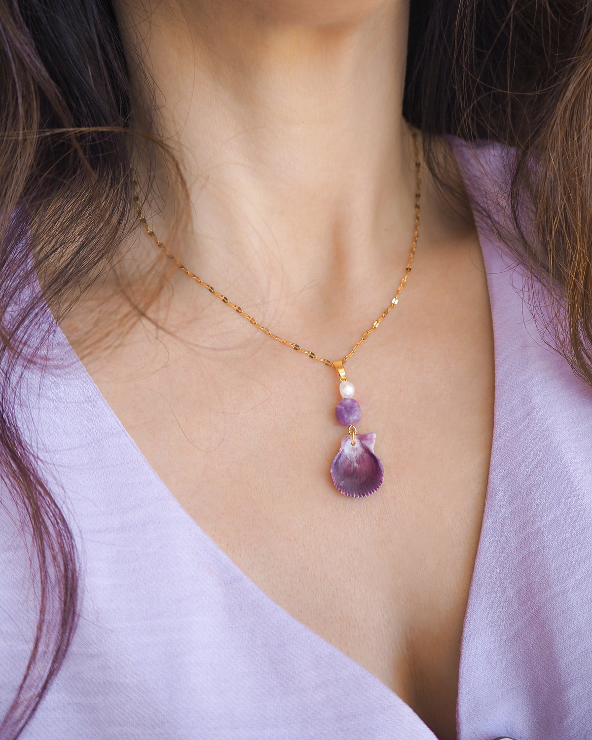 A close-up of a woman wearing a delicate gold chain necklace with a tiny scallop shell, a purple amethyst gemstone bead and a small white pearl bead. The shell is a variegated scallop shell handpicked from the beaches of Portugal and it has a plum bordeaux color.