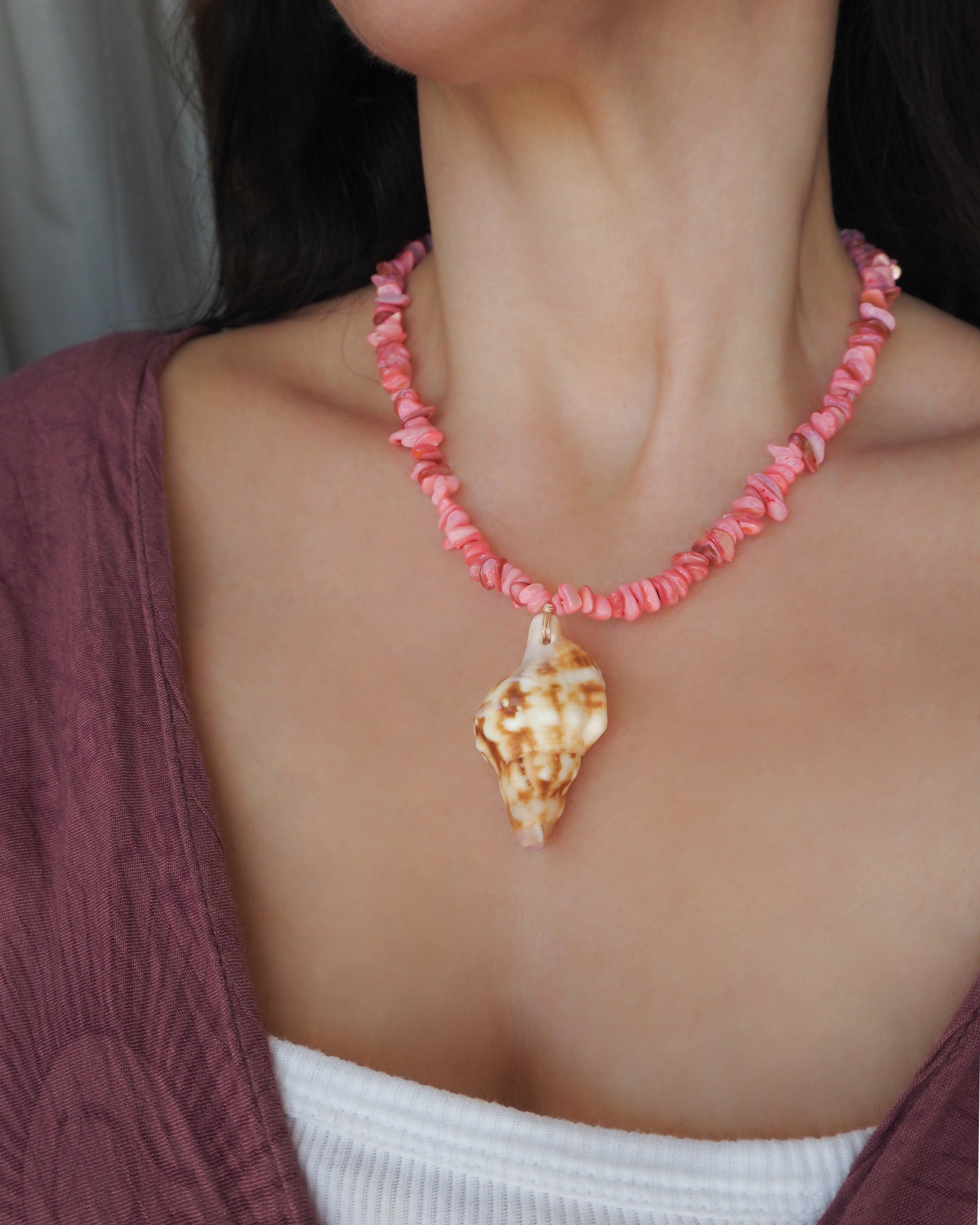 A close-up of a person wearing a necklace made of pink seashell-like beads with a larger spiral shell pendant at the center, styled over a mauve cardigan and white top. Knobbed triton shell necklace with coral pink stones. The shell is conch shaped with brown and white beige pattern. It's a handpicked shell from Portugal. 