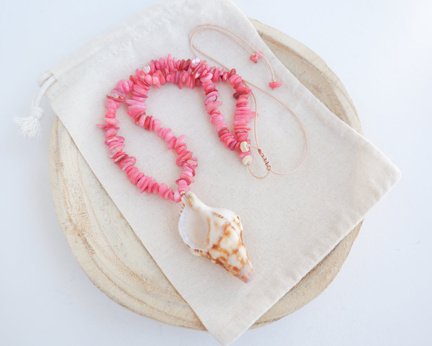 A pink shell bead necklace with a large conch shell pendant, displayed on a cream fabric drawstring pouch over a round wooden tray against a white background. Knobbed triton shell necklace with coral pink stones. The shell is conch shaped with brown and white beige pattern. It's a handpicked shell from Portugal. 