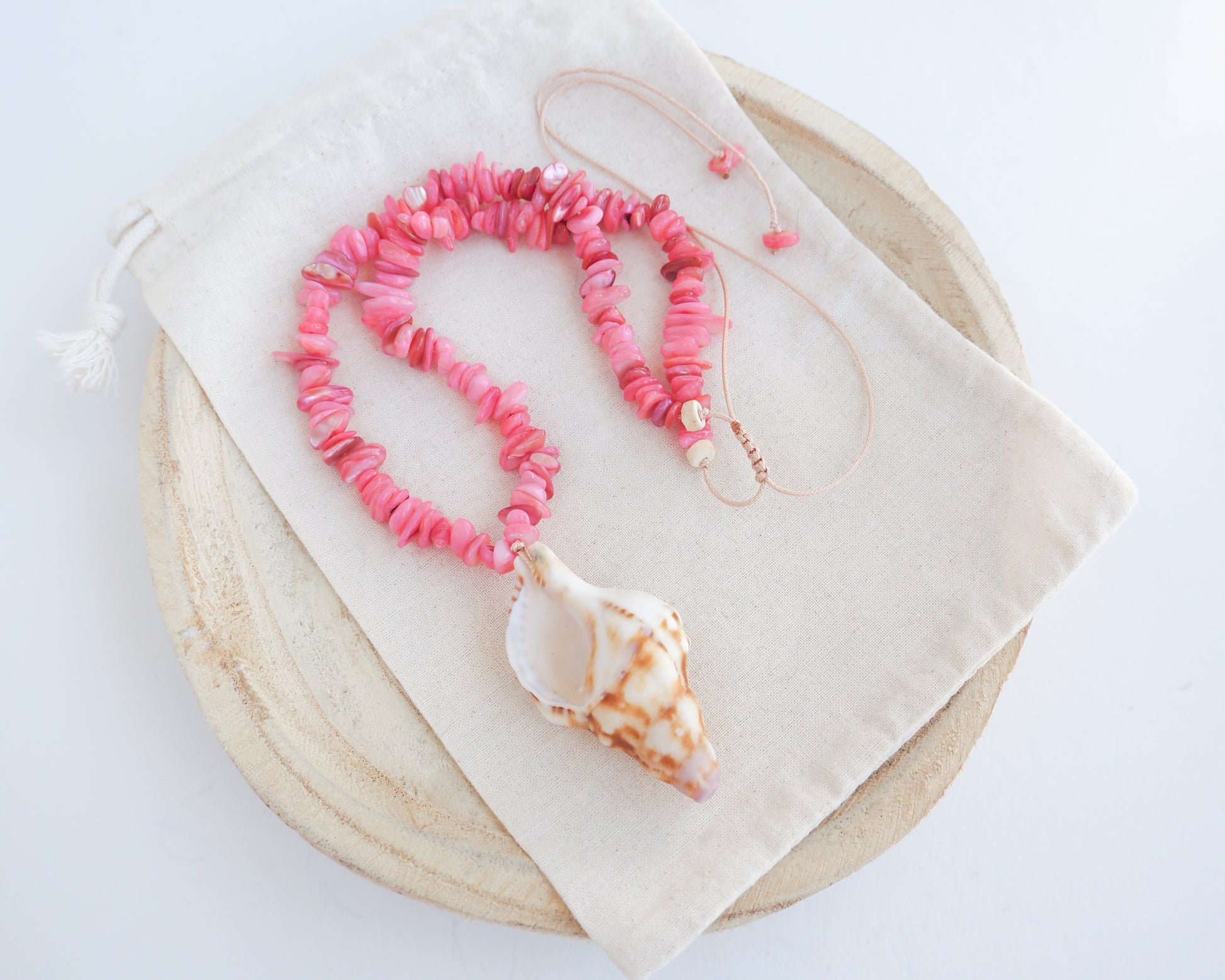 A pink shell bead necklace with a large conch shell pendant, displayed on a cream fabric drawstring pouch over a round wooden tray against a white background. Knobbed triton shell necklace with coral pink stones. The shell is conch shaped with brown and white beige pattern. It's a handpicked shell from Portugal. 