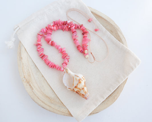A pink shell bead necklace with a large conch shell pendant, displayed on a cream fabric drawstring pouch over a round wooden tray against a white background. Knobbed triton shell necklace with coral pink stones. The shell is conch shaped with brown and white beige pattern. It's a handpicked shell from Portugal. 