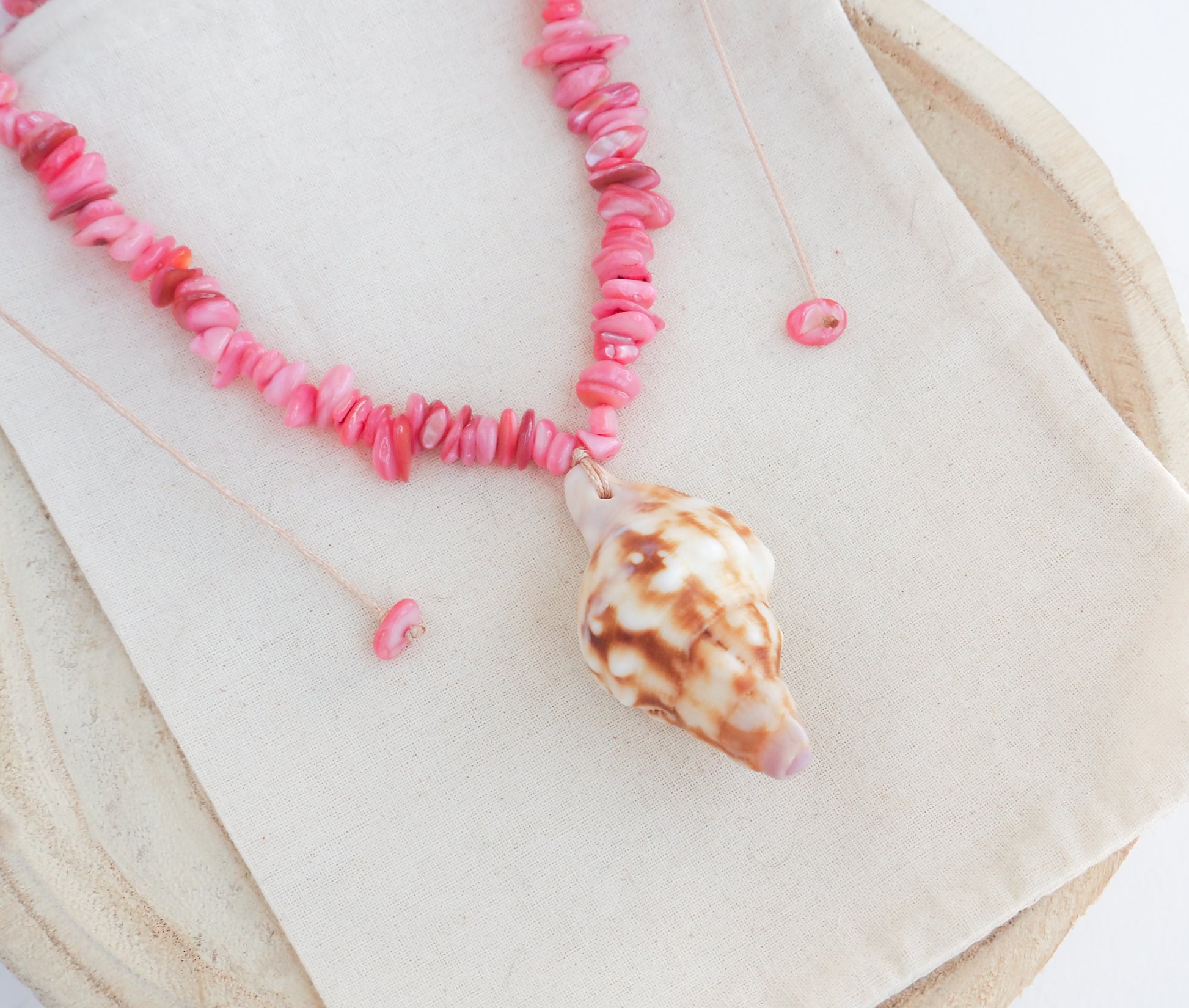 A pink beaded necklace with a conch shell pendant, displayed on a beige fabric surface with a wooden tray background. Knobbed triton shell necklace with coral pink stones. The shell is conch shaped with brown and white beige pattern. It's a handpicked shell from Portugal. 