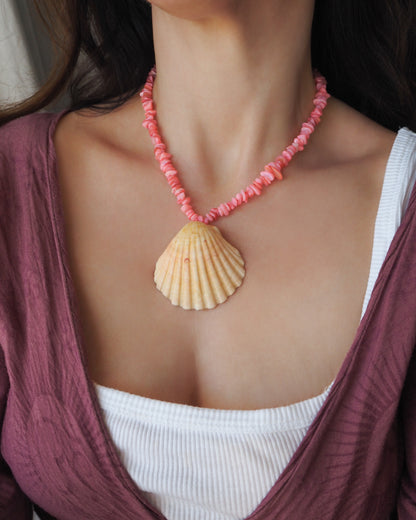 A close-up of a woman wearing a pink beaded necklace with a large beige scallop shell pendant, styled over a white ribbed tank top and a mauve wrap cardigan. A scallop shell necklace with soft coral pink stone beads. The shell is a pilgrim's scallop shell from Portugal and has soft orange coral color. 