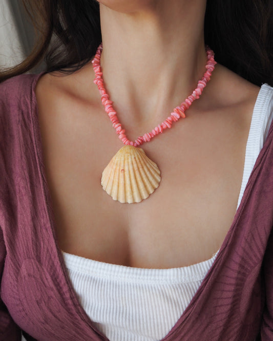 A close-up of a woman wearing a pink beaded necklace with a large beige scallop shell pendant, styled over a white ribbed tank top and a mauve wrap cardigan. A scallop shell necklace with soft coral pink stone beads. The shell is a pilgrim's scallop shell from Portugal and has soft orange coral color. 