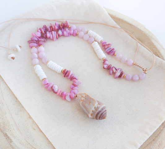 A pink and white beaded necklace with a large seashell pendant, displayed on a beige fabric surface. A necklace with pink and white beads and a large cone shell pendant on a light background. A necklace with a mediterranean cone shell paired with rose quartz gemstone beads, soft pink stones and white shell beads. The shell is a mediterranean cone shell from Portugal and is about 3,5 cm. The color of the shell is brown.
