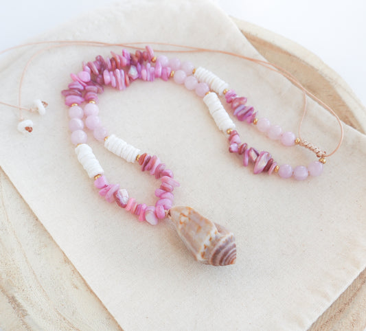 A pink and white beaded necklace with a large seashell pendant, displayed on a beige fabric surface. A necklace with pink and white beads and a large cone shell pendant on a light background. A necklace with a mediterranean cone shell paired with rose quartz gemstone beads, soft pink stones and white shell beads. The shell is a mediterranean cone shell from Portugal and is about 3,5 cm. The color of the shell is brown.