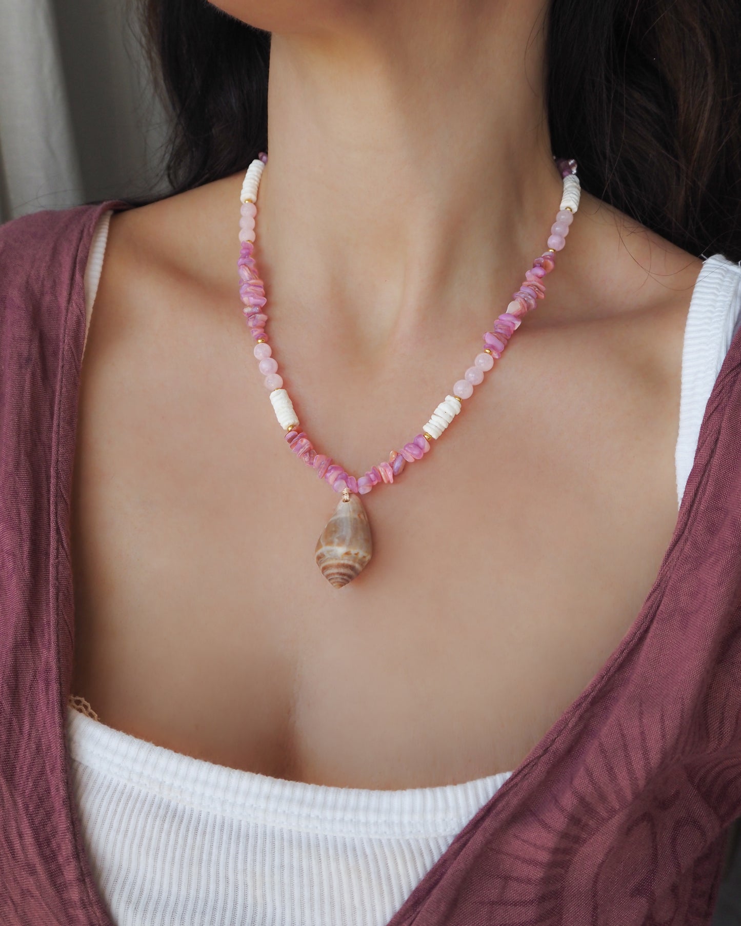 Model wearing a necklace with pink and white beads and a large cone shell pendant on a light background. A necklace with a mediterranean cone shell paired with rose quartz gemstone beads, soft pink stones and white shell beads. The shell is a mediterranean cone shell from Portugal and is about 3,5 cm. The color of the shell is brown.